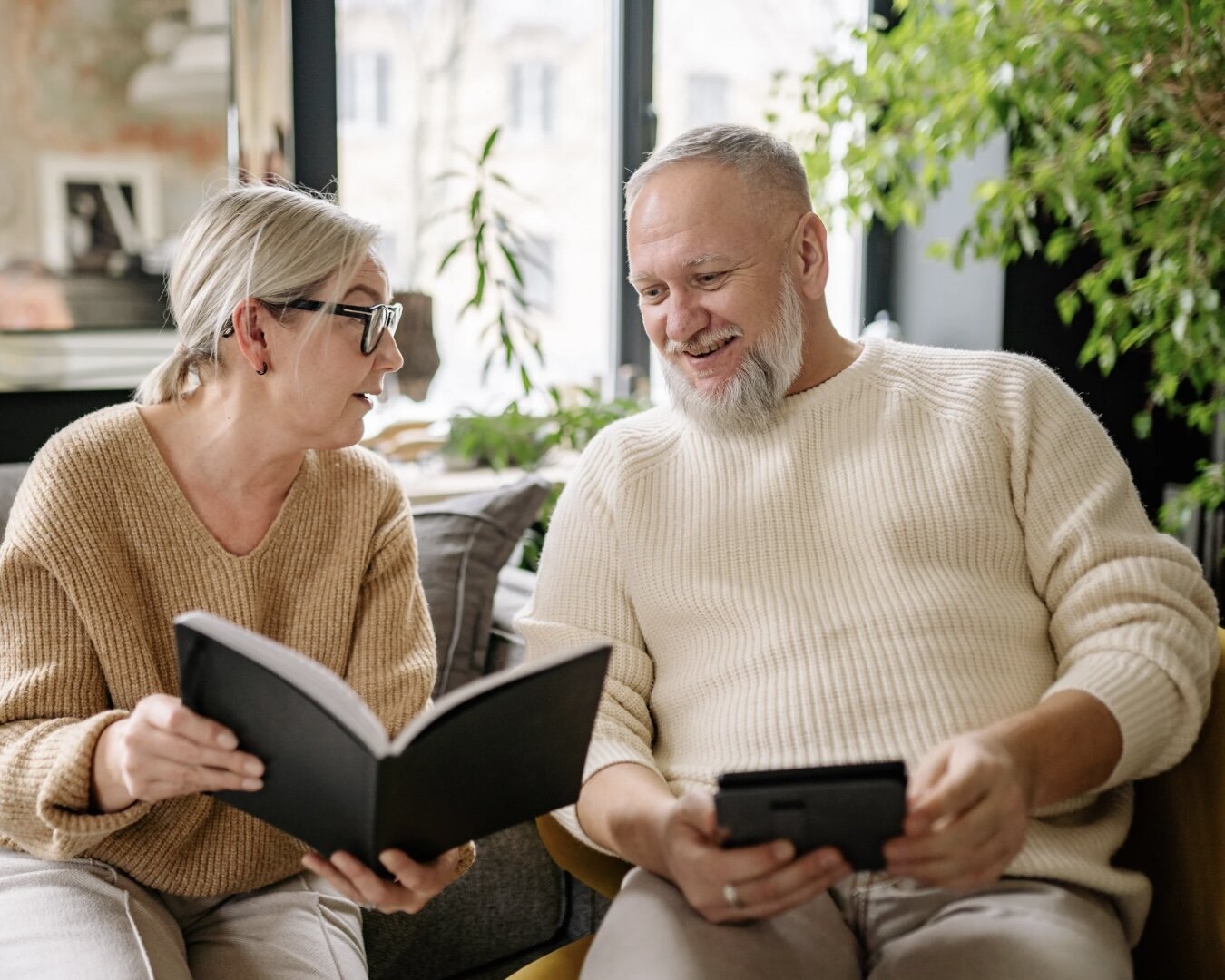 Two thoughtful people seated by a window, leaning in together over a leather portfolio and tablet — partnership over careful, considered conversation